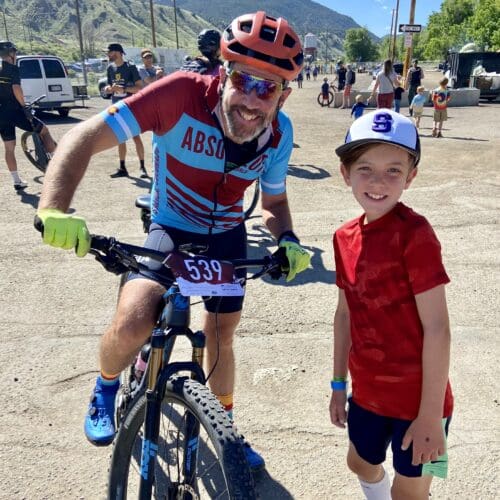 John Witherspoon with his racing shirt and helmet on, on his mountainbike, smiling with a youth standing next to him